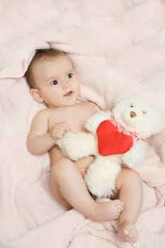 An adorable, charming baby is happily cuddling a soft teddy bear while lying on a cozy pink blanket