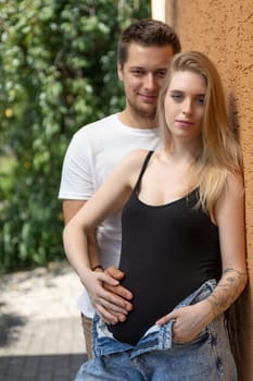 Pregnant young blonde woman is posing with her man leaning against the orange wall.  Vertically.