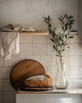 A rustic kitchen features freshly baked bread on a wooden board, with a simple olive branch in a glass vase, creating a warm ambiance.