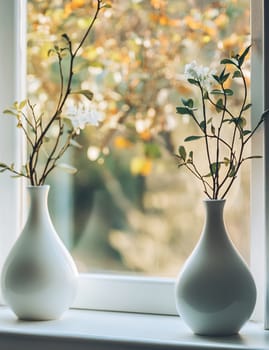 Two white vases hold delicate branches with flowers, illuminated by soft sunlight coming through the window.