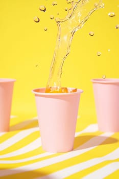 A colorful drink is splashed into a pink cup, surrounded by more cups against a bright yellow background.