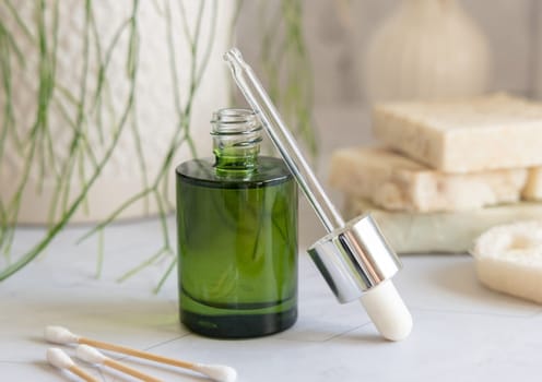 Opened green cosmetic dropper bottle on white marble against green plant, pipette resting on its edge, close up. Skincare or spa concept for beauty and relaxation