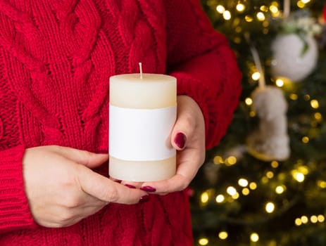 Cream pillar candle with blank label in female hands against Christmas tree lights, mock up, copy space, front view. Yound woman in red sweater keeps candle. Holidays atmosphere