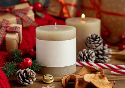 Pillar candles with blank label near red knitted sweater and Christmas decor against lights and wrapped presents, copy space. Warm winter family atmospheric mock up, Holiday time