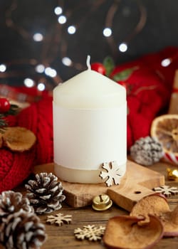 Pillar candle with blank label near red knitted sweater and Christmas decor against lights and wrapped presents, copy space. Warm winter family atmospheric mock up, Holiday time