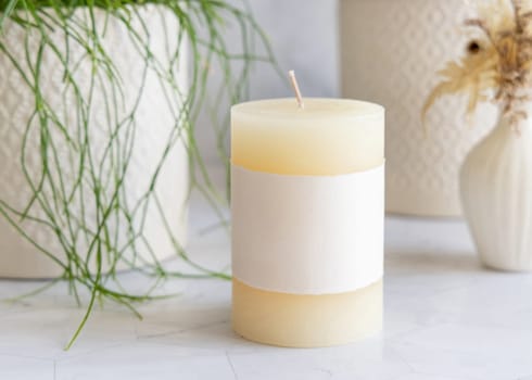 Pillar candle with blank label on marble near potted green plant close up, packaging mockup. Minimal scandinavian home decoration, romantic interior