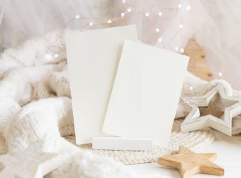 Vertical blank paper cards near white knitted sweater, wooden Christmas decor and lights, copy space. Winter atmospheric mock up, family greeting in rustic or scandinavian style