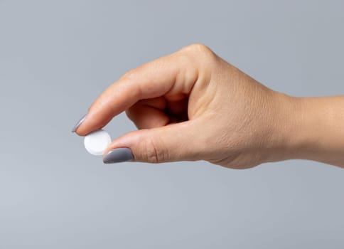 Female hand holding a white pill with fingers closeup against grey background. Healthcare, medical and pharmaceutical concept.