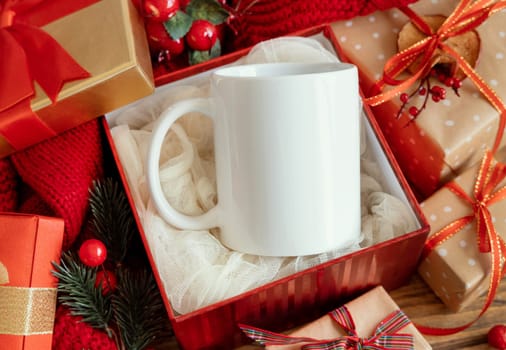White blank coffee mug in gift box near red knitted sweater and Christmas decor against lights and wrapped presents, copy space. Warm winter family atmospheric mock up