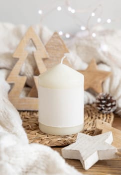 Pillar candle with blank label near white knitted sweater, wooden Christmas decor and lights, copy space. Winter atmospheric mock up in rustic or scandinavian style