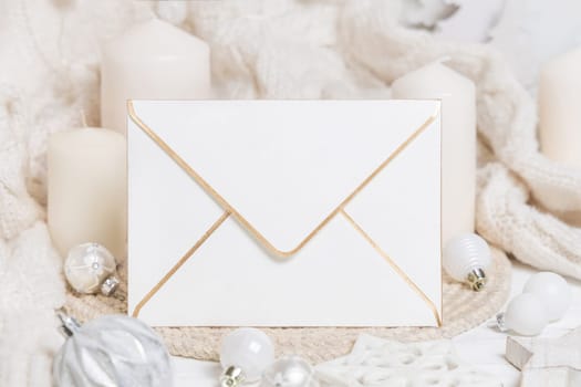 Blank envelope near white knitted sweater, pillar candles, Christmas baubles and decor, copy space. Pastel winter atmospheric mock up for family greeting