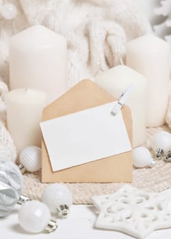 Blank paper card and envelope near white knitted sweater, pillar candles and Christmas baubles and decor, copy space. Winter atmospheric mock up for family greeting