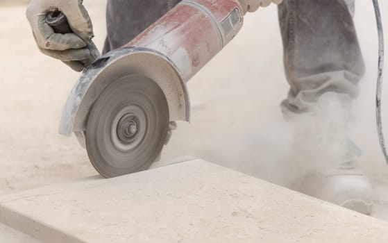 Craftsman cutting marble with circular saw piece against scaffoldings. Men in dusty grey work clothes and white construction helmet during construction process