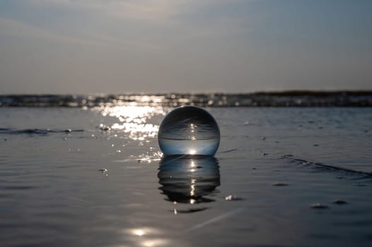 A glass ball lies in the waves on the sandy beach, the sea and the setting sun are reflected in the ball