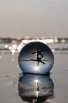 A glass ball lies in the waves on the sandy beach, the ball reflects the sea, a starfish and the setting sun