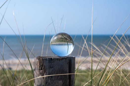 Glass ball lies on a wooden post, with beach grass , the sea, the sun and the blue sky are reflected in the ball