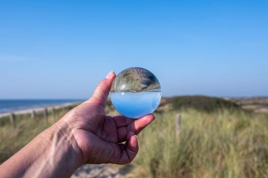 A human hand holds a glass ball, in the ball a sand dune, a footpath, blue sky and the sea are reflected