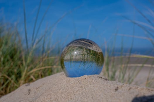A glass ball lies in the sand of a dune by the sea, the dune grass and the blue sky are reflected in the ball