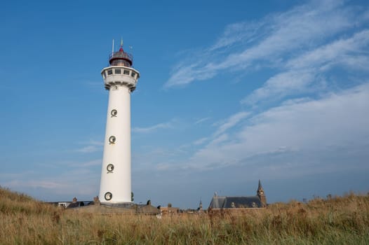 The approx. 28 m high white lighthouse J.C.J. van Speijk in Egmond aan Zee, North Holland, Netherlands with the Catholic St. Agnes Church, beach grass and sand dunes