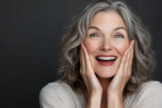 Cheerful senior woman with gray hair smiling widely, hands on cheeks, showing delight and happiness.