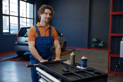 Young man mechanic working at car repair service workshop. Male technician pushing trolley cat with tools and supplies for auto maintenance and tuning