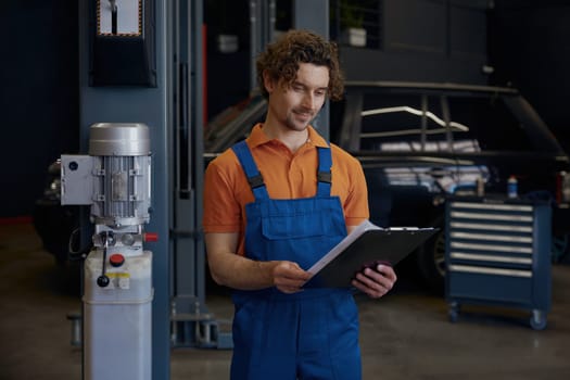 Young friendly smiling male worker with clipboard wearing coveralls standing over car repair service station