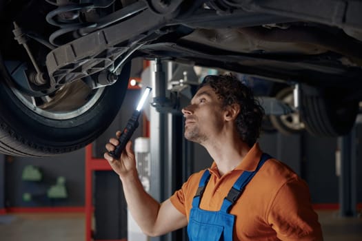 Auto mechanic with flashlight examining brake pads of lifted car nd fixing problem at garage workshop