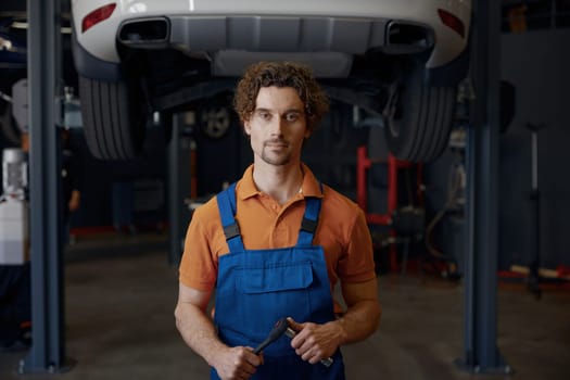 Serious and concentrated man car mechanic holding wrench tool looking at bottom of lifted automobile at repair service station
