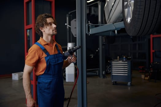 Male auto mechanic using impact wrench during tires replacement standing underneath of car at garage workshop
