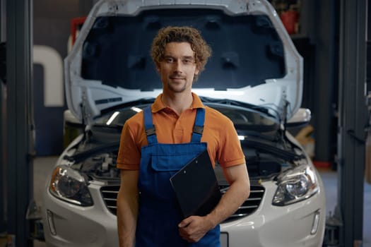 Portrait of young male auto mechanic holding clipboard standing over car with opened hood on repair workshop indoor background