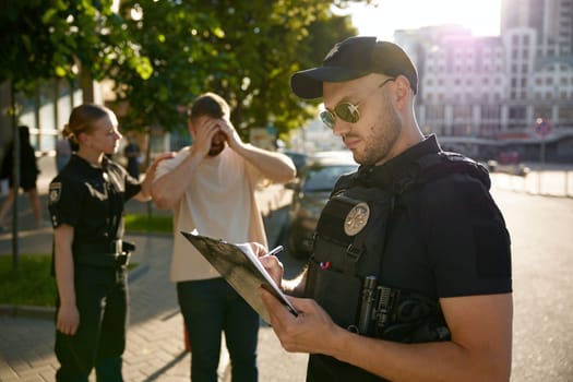 Policeman filling out offense form, while policewoman calming down upset male citizen victim after unfortunate incident on city street
