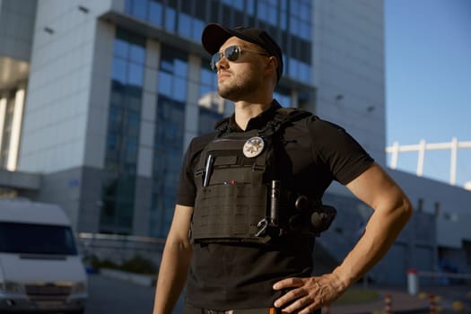 Confident male patrol police officer wearing uniform and eyeglasses standing outdoors against police station. City street security monitoring