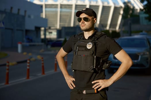 Portrait of confident male patrol police officer wearing uniform and eyeglasses standing outdoors. City street security monitoring