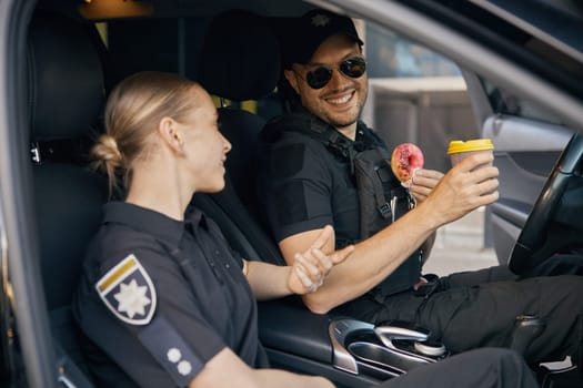 Patrol police officers diverse team sitting in car having coffee break time at work. Female colleague treating coworker with donut and hot aroma drink