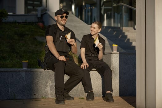 Smiling male and female patrol police officers enjoying lunch break. Policeman and policewoman resting outdoor, drinking coffee, eating sandwich and having friendly conversation