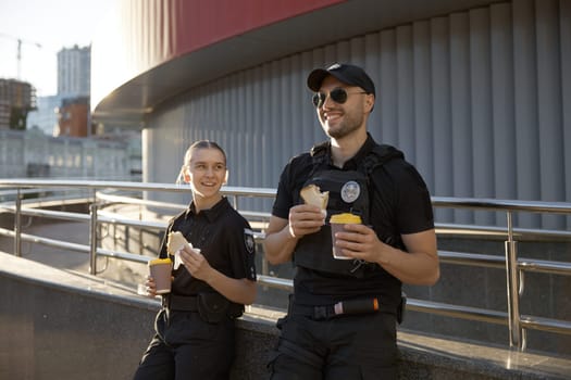 Male and female patrol police officers enjoying lunch break. Policeman and policewoman resting outdoor, drinking coffee, eating sandwich and having friendly conversation