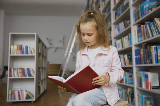 Pretty little girl child reading paper book while spending time in public library for children. Preschool female kid sitting on bench front of bookcase with shelves full of literature