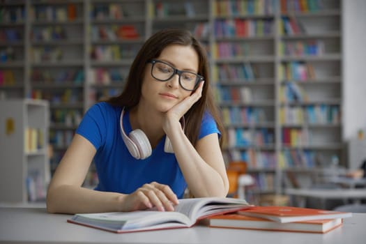 Dreamy young smart woman reading novel book or educational literature sitting at table over bookshelves background in library