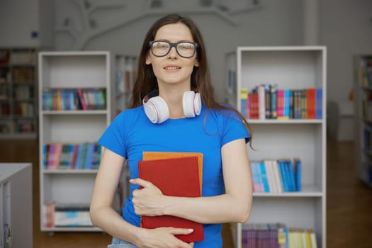 Portrait of young teenage student or smart literature fan wearing eyeglasses and headphones, holding paper books looking at camera standing over public library bookshelves on background