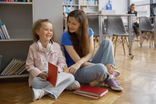 Happy family in library or bookstore. Cheerful mother and joyful daughter sitting on floor spending time together reading funny paper book enjoying educational leisure activity on weekend