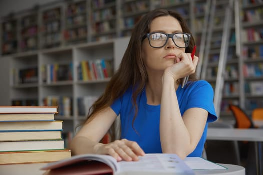 Smart pensive young woman wearing eyeglasses feeling dreamy and thoughtful looking through window while spending time in library. Preparation for exam, self-education, research work at bookstore