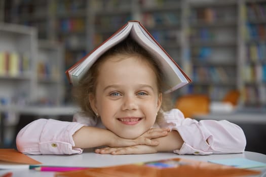 Portrait of cute cheerful happy girl child with book on head sitting at table over library background