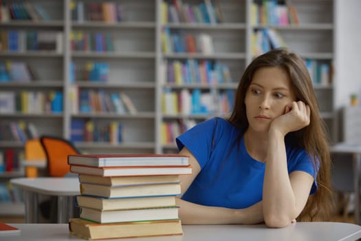 Shocked young woman student looking at huge heap of books on table while preparing for exam in library or shopping for education process in bookstore
