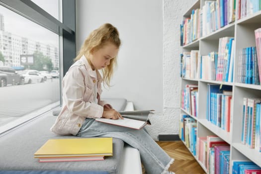 Pretty little girl child reading paper book while spending time in public library for children. Preschool female kid sitting on bench front of bookcase with shelves full of literature