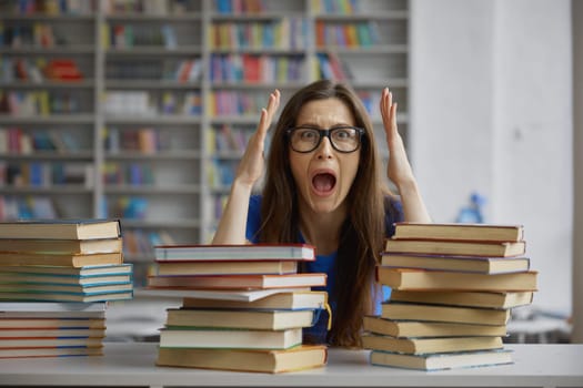 Upset young woman feeling mad and crazy screaming and touching head while sitting front of huge stack of book in library