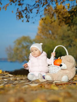 A kid on a picnic with a basket and a pumpkin plays on the riverbank with a teddy bear among colorful autumn leaves