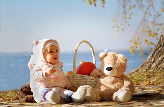 A baby on a picnic with a soft teddy bear and a basket with a pumpkin on the background of the sea