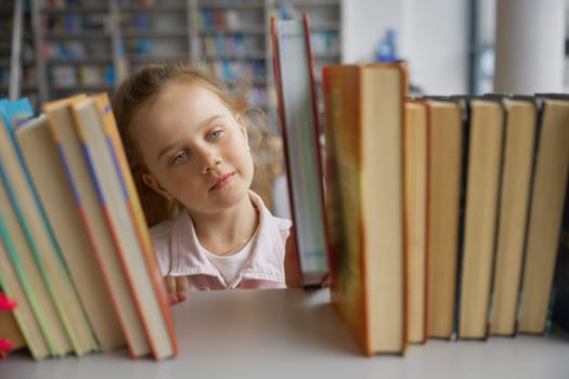 Curious little girl child choosing favorite book from library bookshelf. Cute small reader feeling love for reading