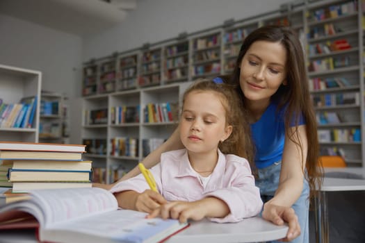 Loving mother assisting adorable little daughter to do homework in library. Cute shot with happy mom and child together over bookshelves background
