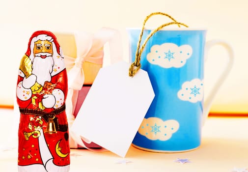 Christmas greeting card. Large blue white clouds winter mug with empty copy space tag, a cheerful wrapped chocolate Santa Claus and giftboxes on table over bright background.